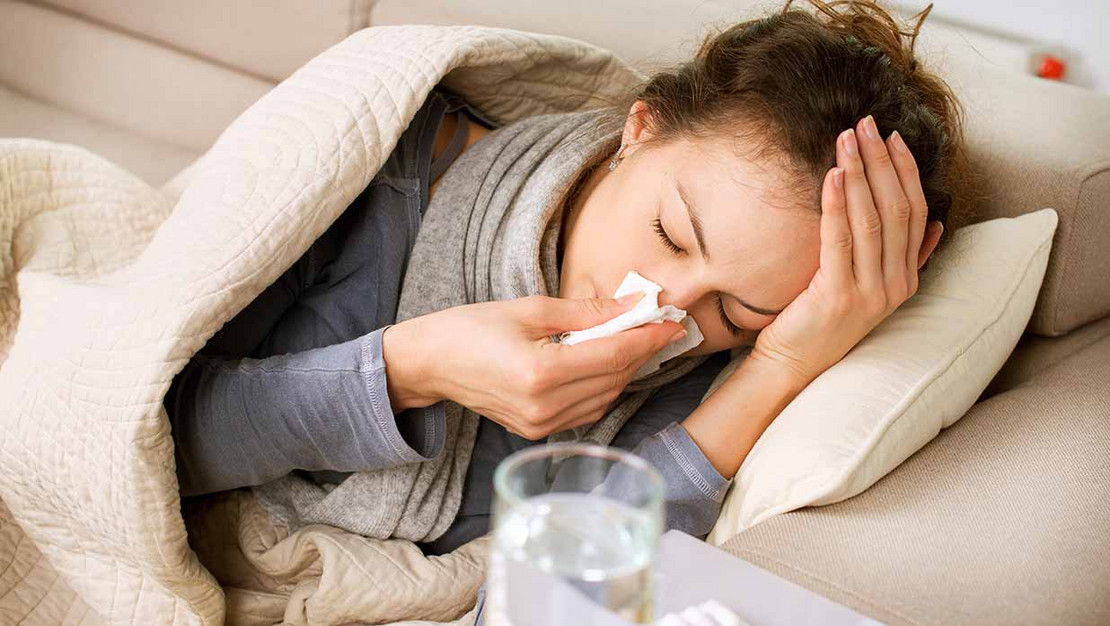 Eine Frau liegt sehr erkältet und in eine Decke eingemummelt auf ihrem Sofa, vor ihr liegen Medikamente.