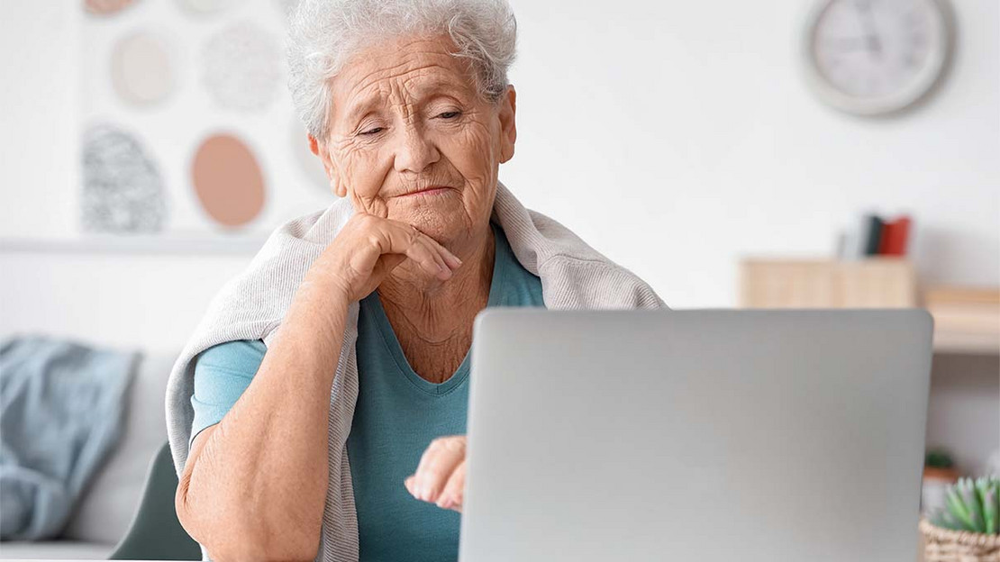 Ältere Frau sitzt vor einem Laptop und blickt skeptisch auf den Bildschirm. 