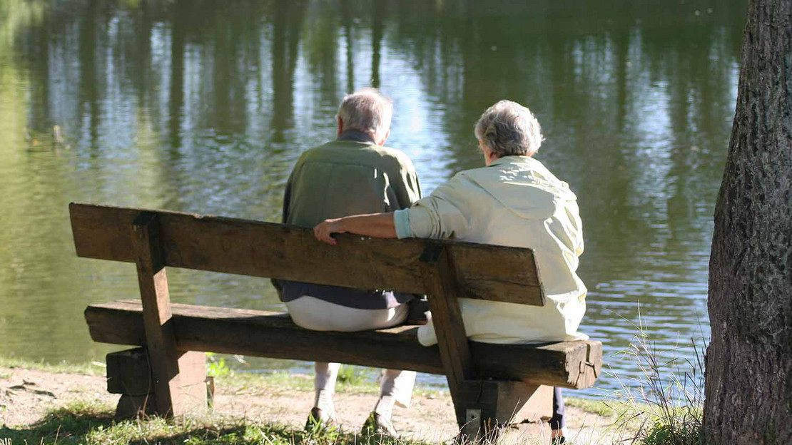 Zwei ältere Personen sitzen mit Jacken auf einer Bank und blicken auf eine Wasserfläche.